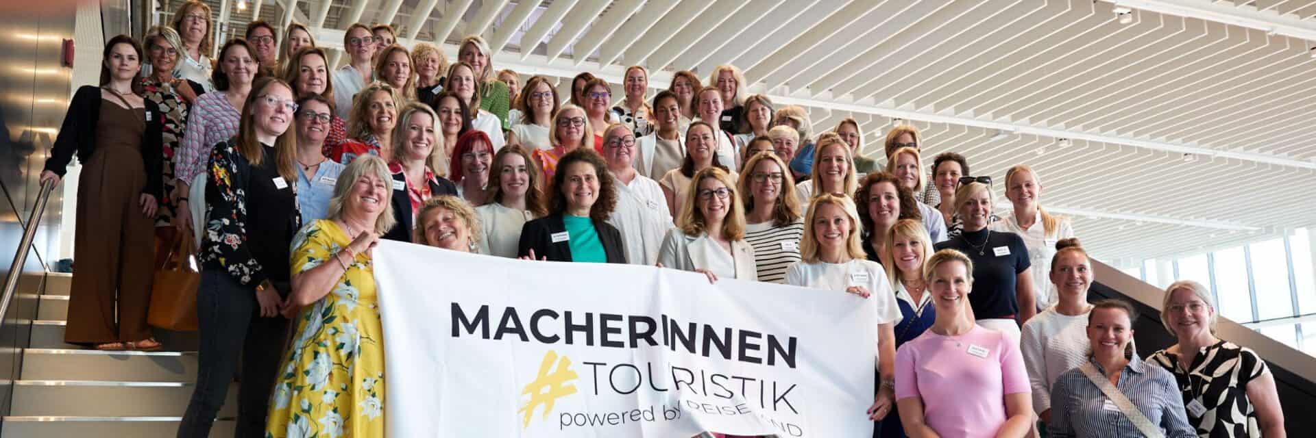 macherinnen-touristik Gruppenszene mit Frauen, die auf einer Treppe stehen und ein Banner mit dem Schriftzug 'MACHERINNEN TOURISTIK' halten.