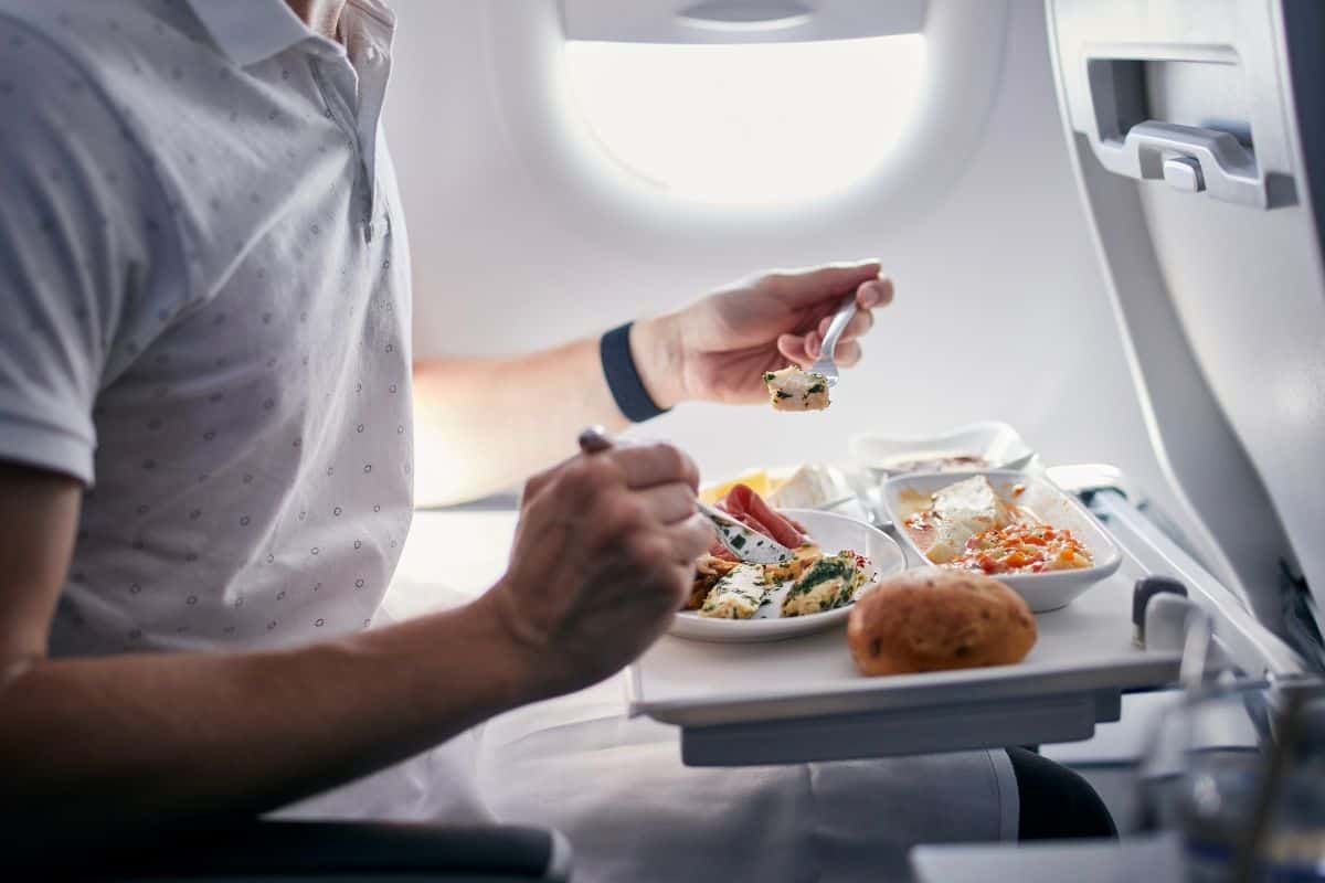 Ein Passagier in einem Flugzeug genießt eine Mahlzeit auf einem Tablett mit verschiedenen Speisen und einem Brötchen, während Licht durch das Fenster fällt.