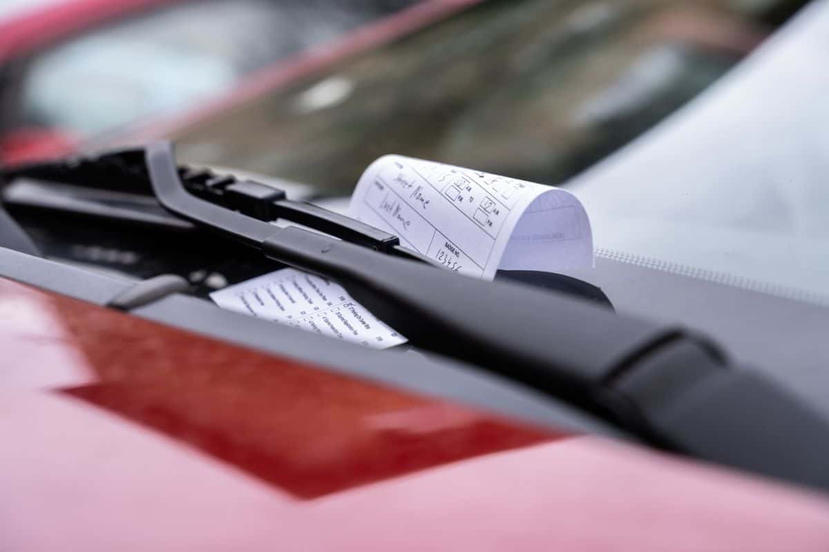 Nahaufnahme eines Parktickets, das unter dem Scheibenwischer eines roten Autos steckt, mit unscharfem Hintergrund.