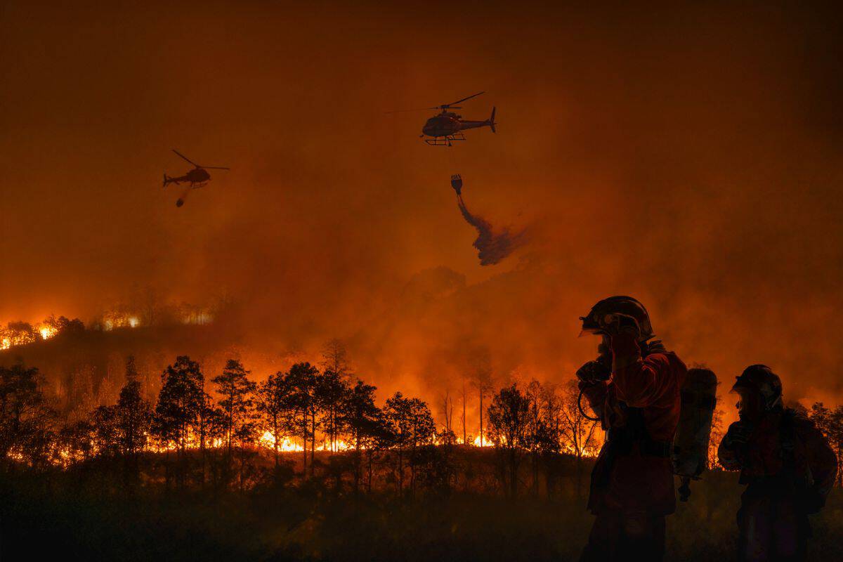Feuerwehrleute stehen vor einem großen Waldbrand, während Hubschrauber Wasser abwerfen. Die Szenerie ist von Rauch und Flammen geprägt.