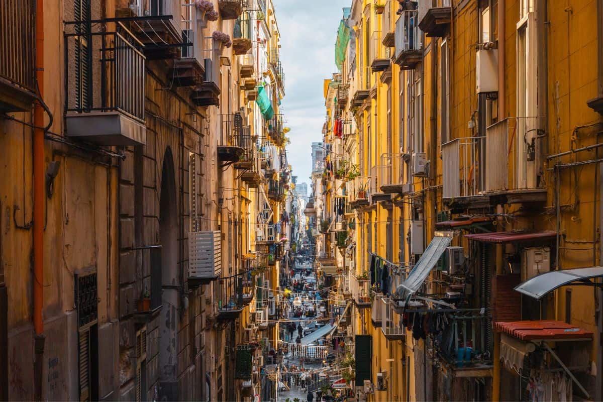 Enge Gasse in einer Stadt mit gelben Gebäuden, Balkonen und Wäsche, die zum Trocknen hängt, unter einem bewölkten Himmel.