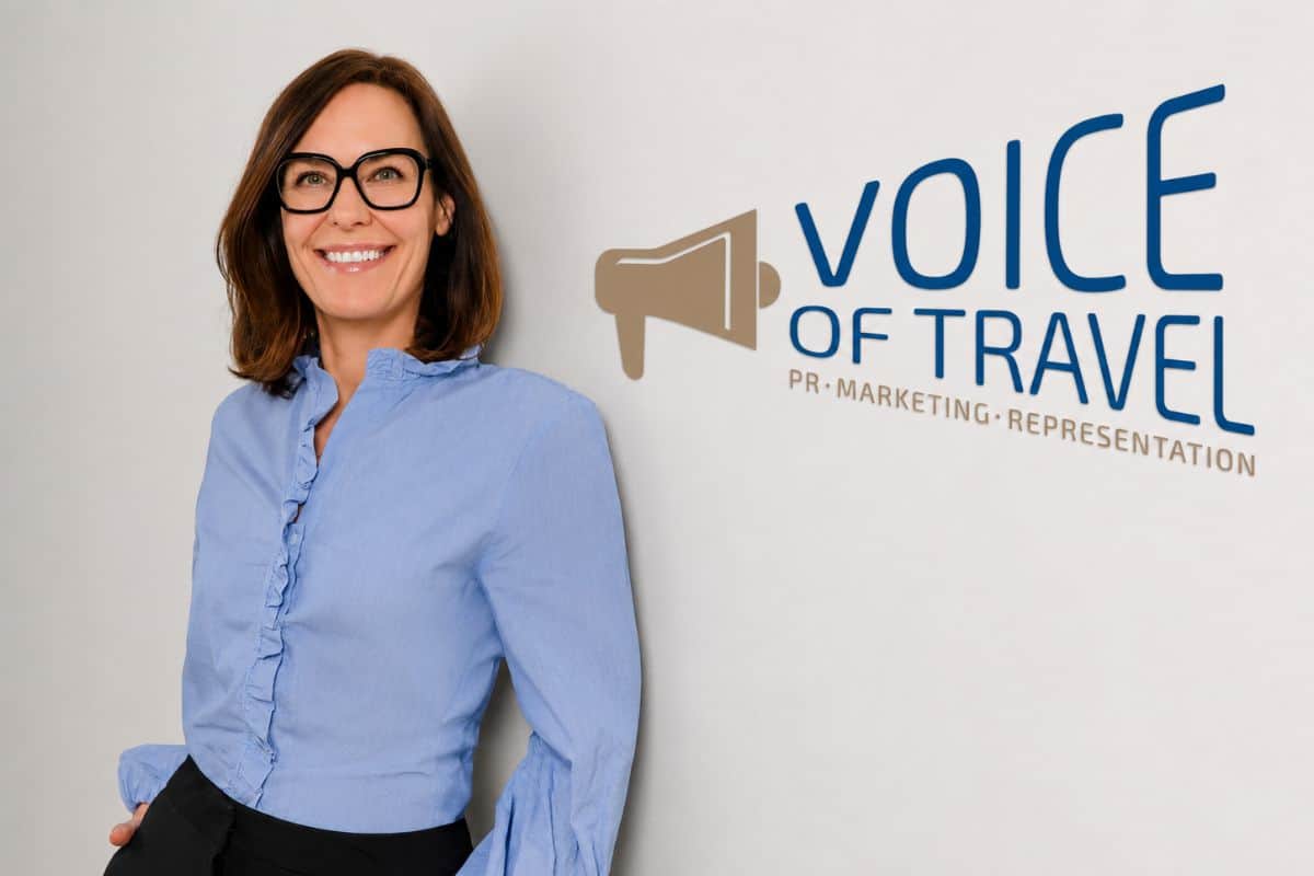 Eine lächelnde Frau mit braunem Haar und Brille trägt eine hellblaue Bluse und steht vor einer weißen Wand mit dem Logo von VOICE OF TRAVEL PR MARKETING.