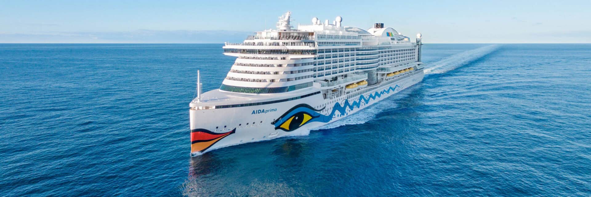 Ein modernes Kreuzfahrtschiff fährt durch ruhige, blaue Gewässer unter einem klaren Himmel. Die Schiffsoberfläche reflektiert das Sonnenlicht.
