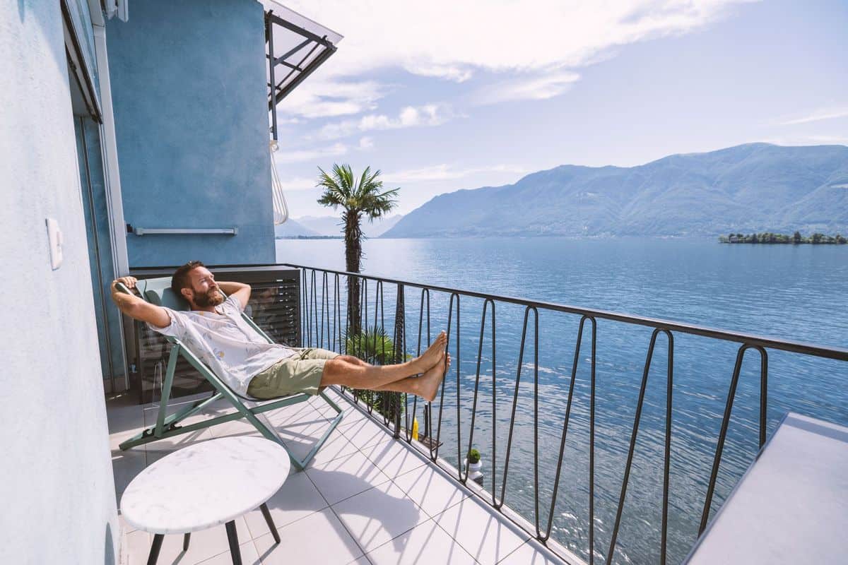 Ein Mann entspannt auf einem Balkon mit Blick auf einen See und Berge, umgeben von einer Palmensilhouette und einem hellen Himmel.