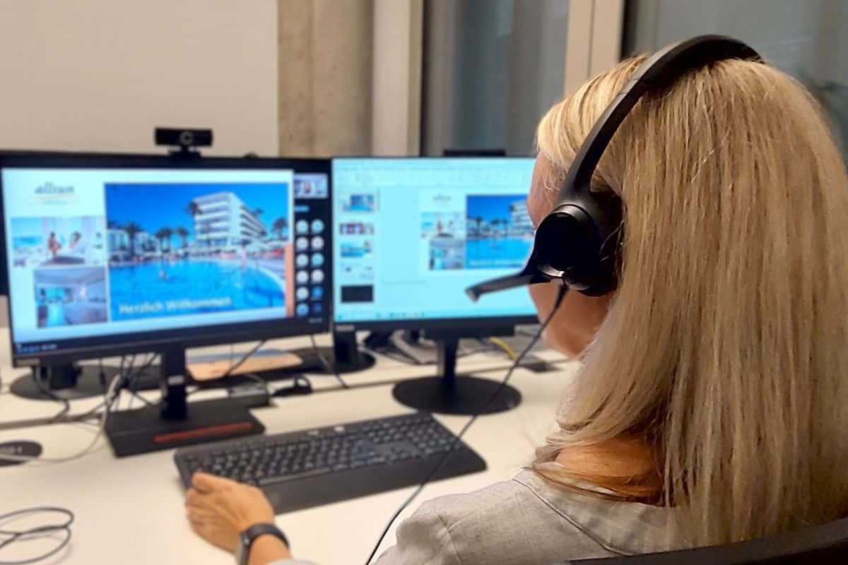 Eine blonde Frau mit Headset sitzt vor zwei Computerbildschirmen an einem Schreibtisch und bedient eine Tastatur. Auf den Monitoren sind Hotelbilder zu sehen.