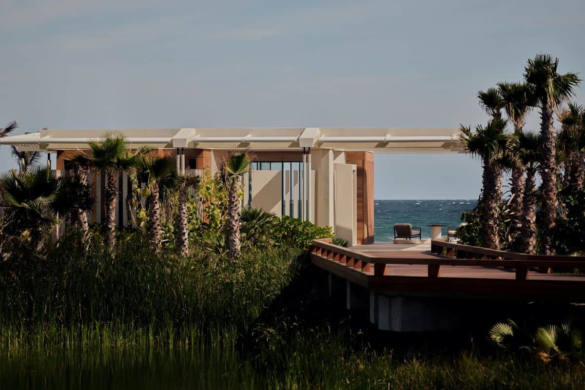 Moderne Architektur eines Gebäudes umgeben von Palmen und üppigem Grün, mit Blick auf das Meer und einer Holzterrasse im Vordergrund.