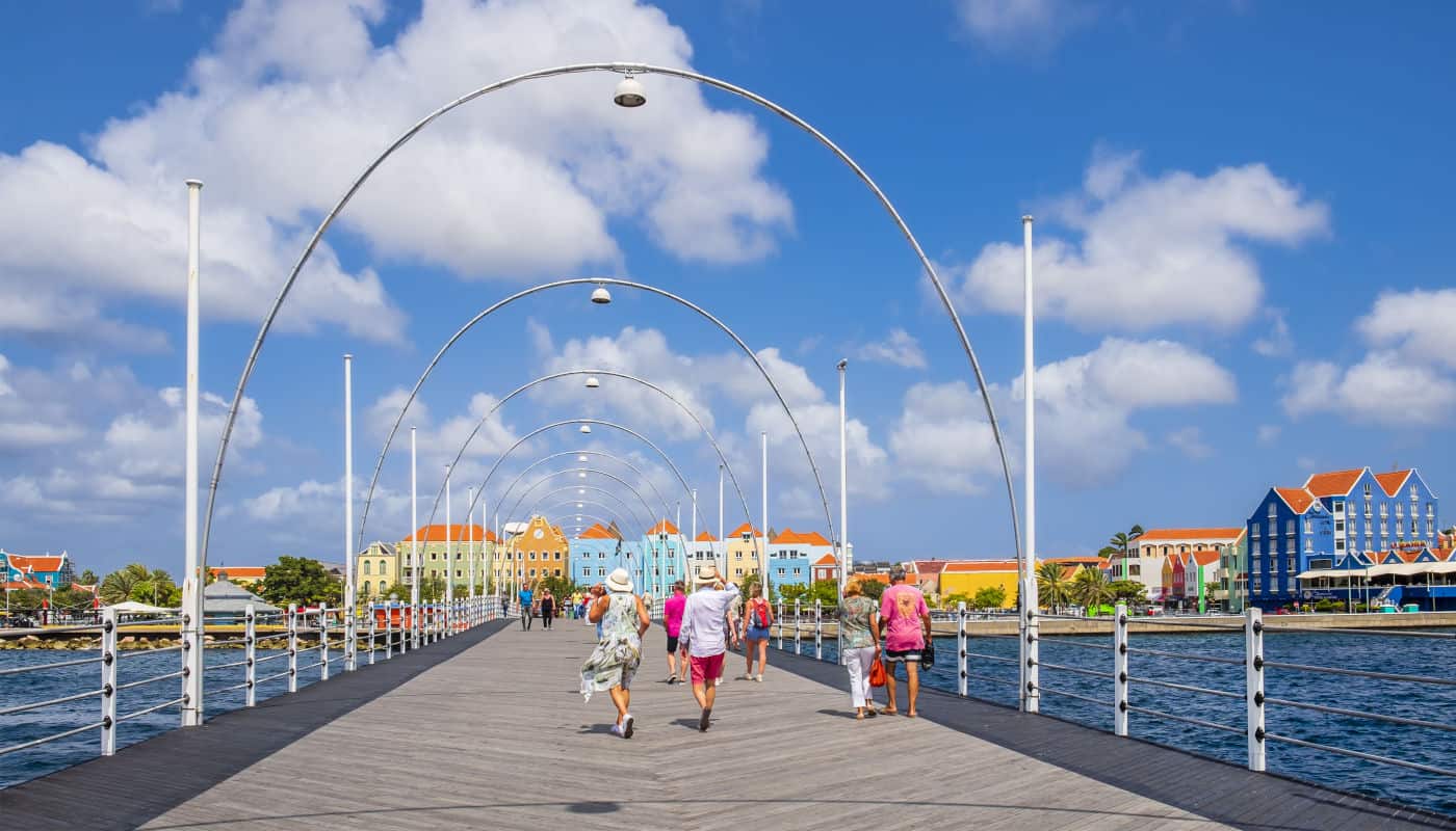 Blick auf eine Holzpromenade mit mehreren Personen, die in Richtung bunter Gebäude am Wasser spazieren.