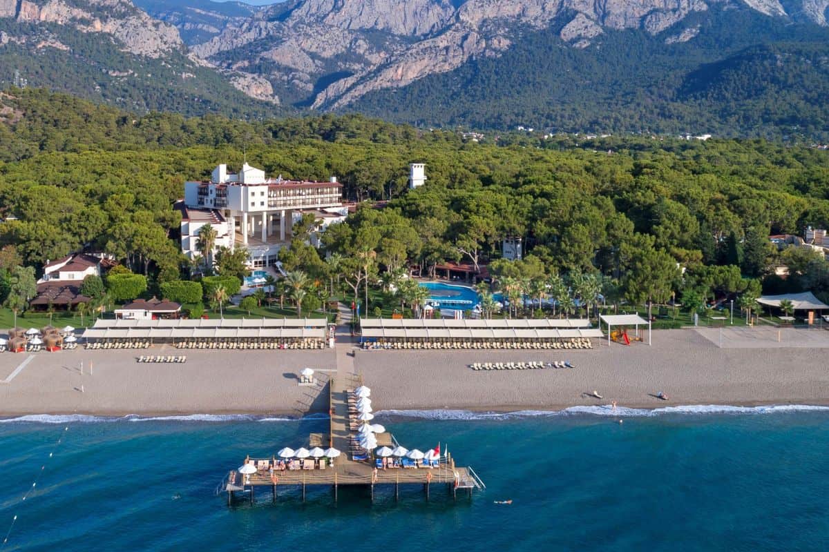Luftaufnahme eines Strandes mit Liegen, einem Steg und einem Hotel im Hintergrund, umgeben von grünen Bäumen und Bergen.