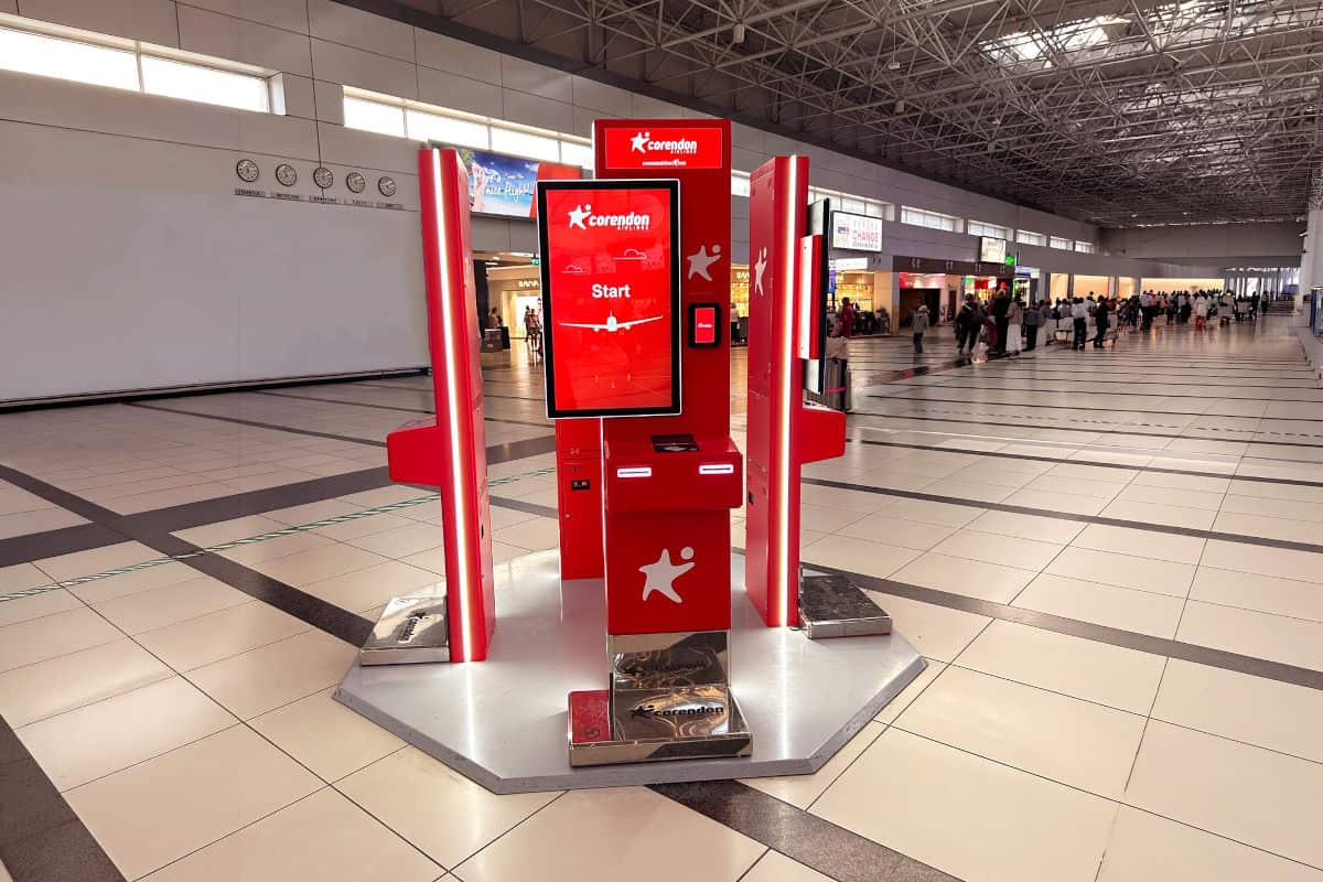 Ein roter Check-in-Automat von Corendon Airlines in einem modernen Flughafen mit hellen Fliesen und einem belebten Hintergrund.