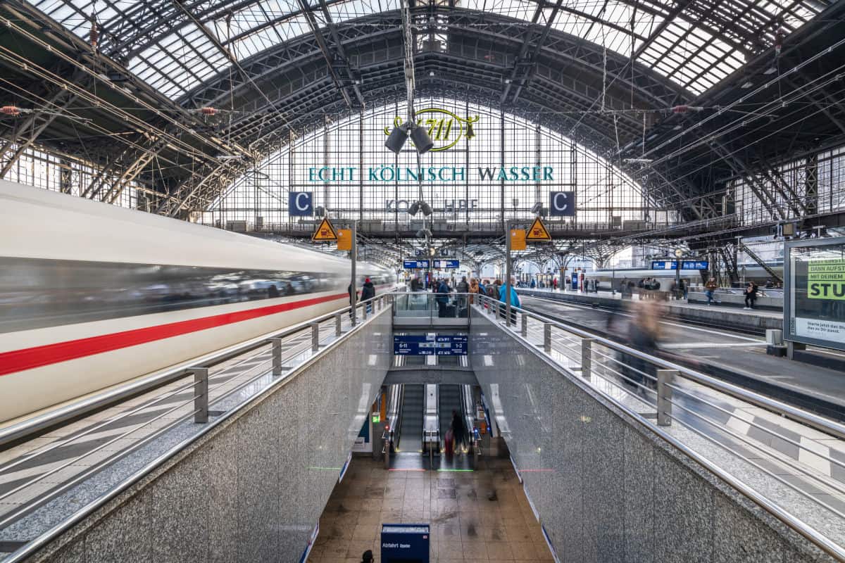 Blick auf den Kölner Hauptbahnhof mit Reisenden und einem schnell vorbeifahrenden Zug unter dem beeindruckenden Glasdach.