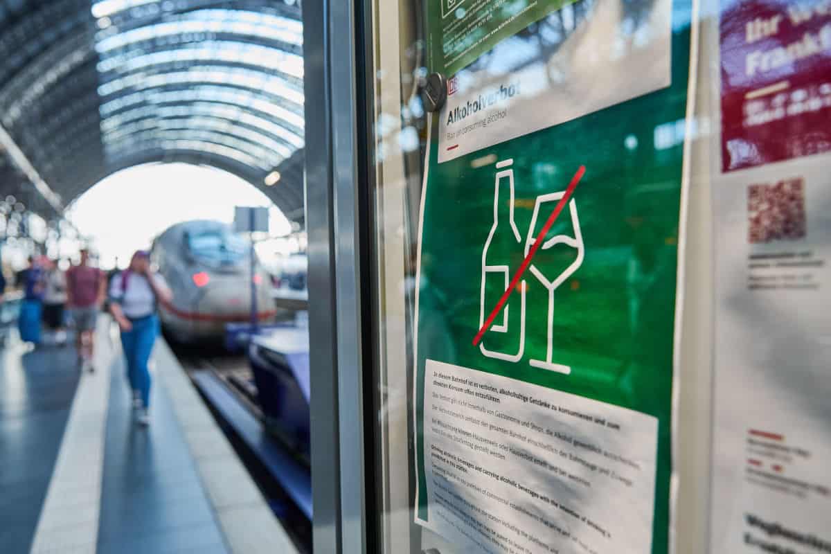 Ein Hinweisschild mit einem Alkoholverbot in einem Bahnhof, im Hintergrund ist unscharf ein einfahrender ICE an einem Bahnsteig mit Passanten zu erkennen.