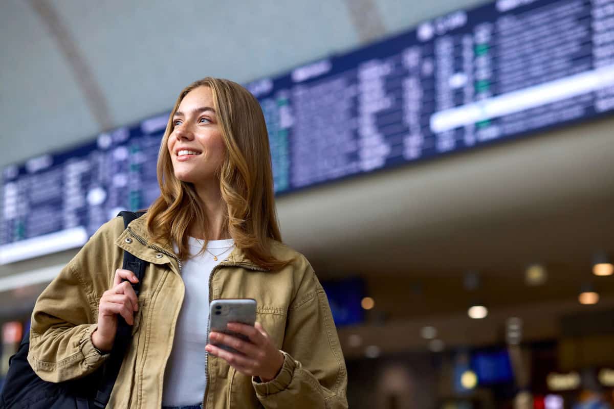 Junge Frau mit Smartphone und Umhängetasche steht in einem Flughafen, im Hintergrund sind Anzeigetafeln mit Fluginformationen sichtbar.