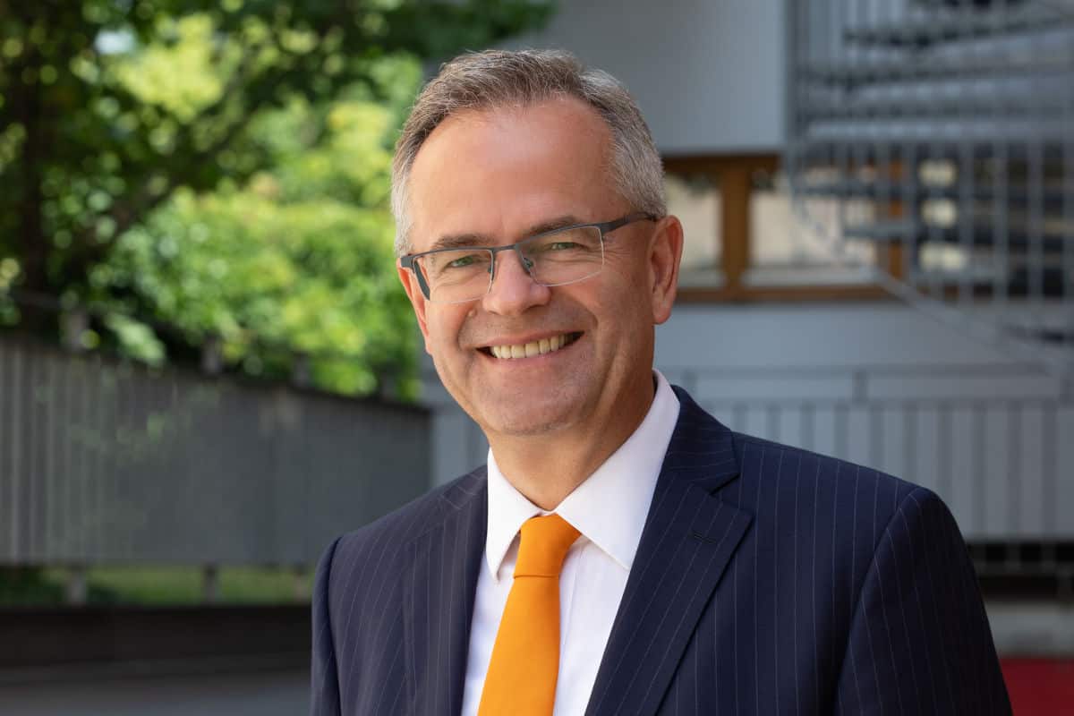 Ein lächelnder Mann mittleren Alters mit Brille trägt ein dunkelblaues Nadelstreifensakko, ein weißes Hemd und eine leuchtend orangefarbene Krawatte im Freien.