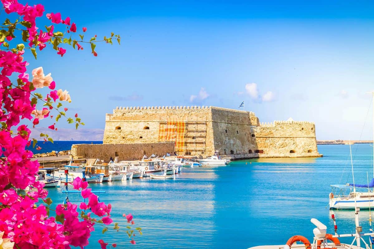 Blick auf eine historische Festung im Wasser, umgeben von bunten Booten und blühenden Pflanzen vor klarem, blauem Himmel.