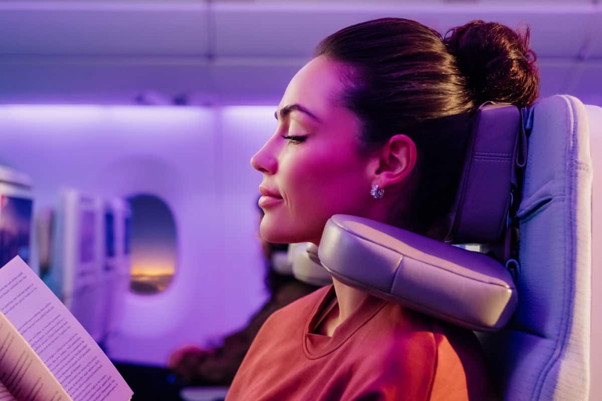Eine Frau mit hochgesteckten Haaren sitzt in einem Flugzeugsitz mit Kopfstütze, liest ein Buch und ist in violettes Licht getaucht, im Hintergrund ein Fenster.