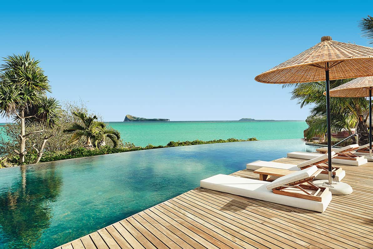 Ein luxuriöser Infinity-Pool mit Blick auf das türkisfarbene Meer, umgeben von tropischen Pflanzen und Sonnenschirmen auf einer Holzterrasse.