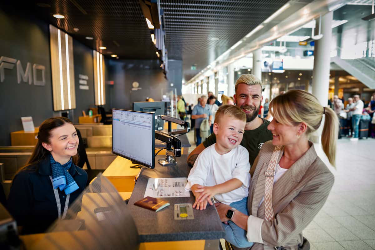 Familie am Check-in-Schalter eines Flughafens, lächelnd mit einem Kind, während eine Mitarbeiterin am Computer arbeitet.