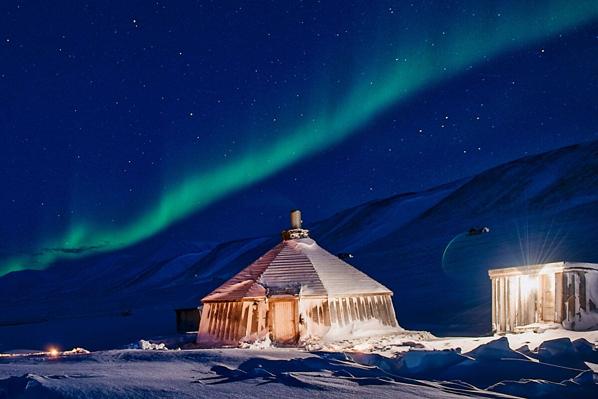 Ein traditionelles, rundes Holzhaus im Schnee, beleuchtet von einem sanften Licht, während der Nachthimmel mit dem Nordlicht erleuchtet wird.