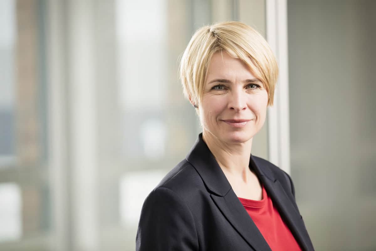 Porträt einer Frau mit blonden, kurzen Haaren, die in einem modernen Büro steht und in die Kamera lächelt.