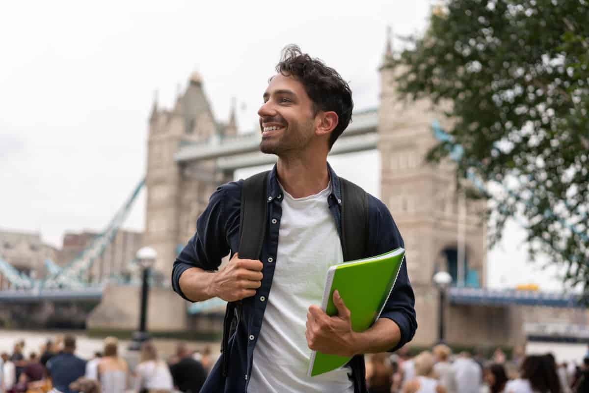 Ein junger Mann mit Rucksack und grünem Notizbuch lächelt vor der Tower Bridge in London, während im Hintergrund unscharf weitere Personen zu sehen sind.