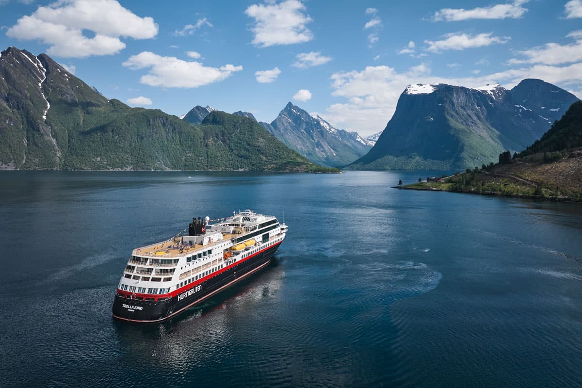 Ein Kreuzfahrtschiff fährt durch einen Fjord, umgeben von majestätischen Bergen und klarem Wasser unter einem blauen Himmel mit wenigen Wolken.