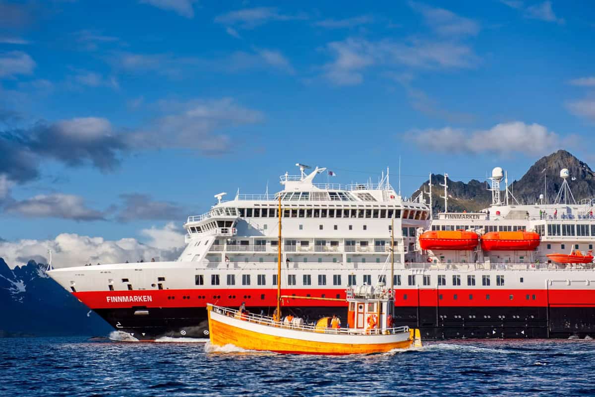Ein großes Kreuzfahrtschiff mit dem Namen Finnmarken fährt durch ruhiges Wasser, während ein kleineres, gelbes Boot in der Nähe navigiert. Im Hintergrund sind Berge sichtbar.
