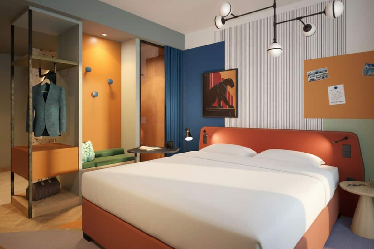 Ein modernes Hotelzimmer mit einem Doppelbett, orangefarbenem Kopfteil, offener Garderobe, einer Sitzbank und grafischer Wandgestaltung in bunten Farben.