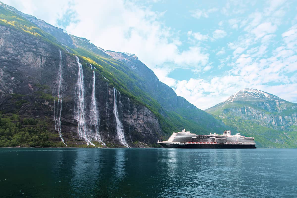 Ein großes Kreuzfahrtschiff liegt ruhig in einem Fjord, umgeben von steilen, bewaldeten Bergen und Wasserfällen, unter einem klaren Himmel.