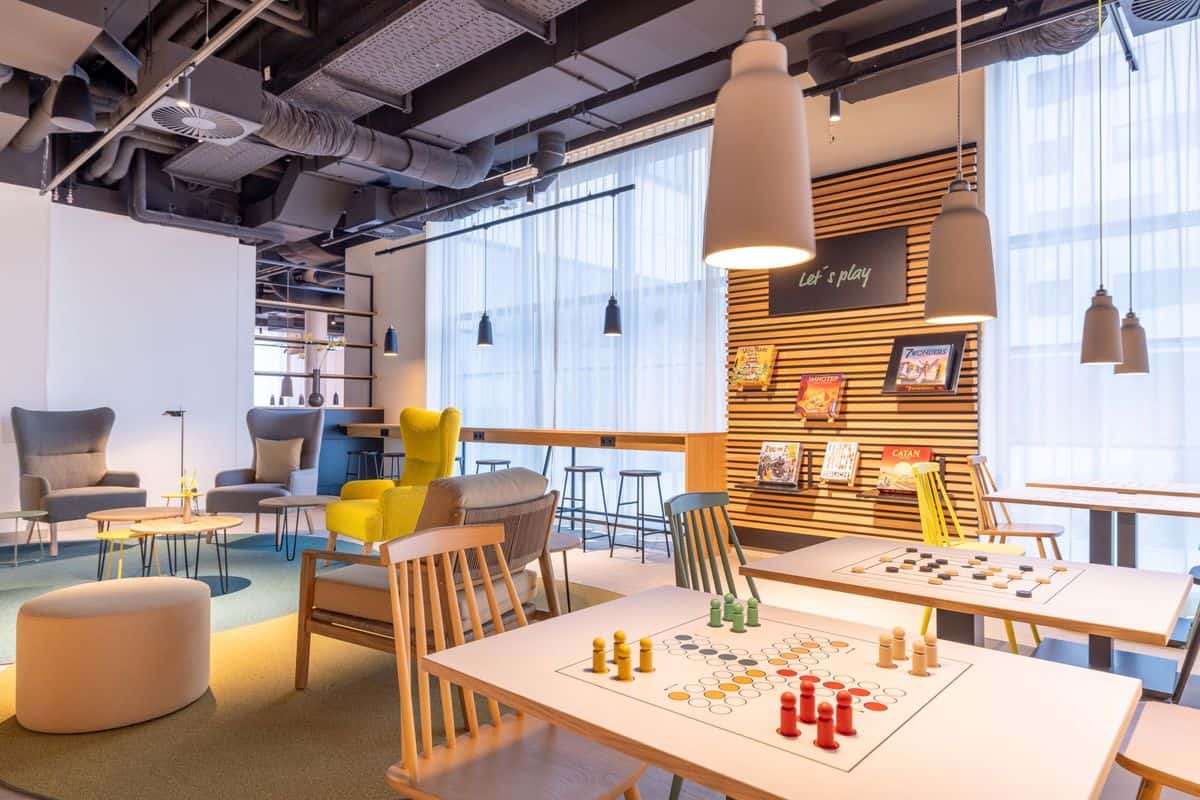 Moderne Lounge mit gemütlichen Sitzgelegenheiten, Spieltischen und einer Bücherwand, die Gesellschaftsspiele präsentiert, in einem hellen Raum.