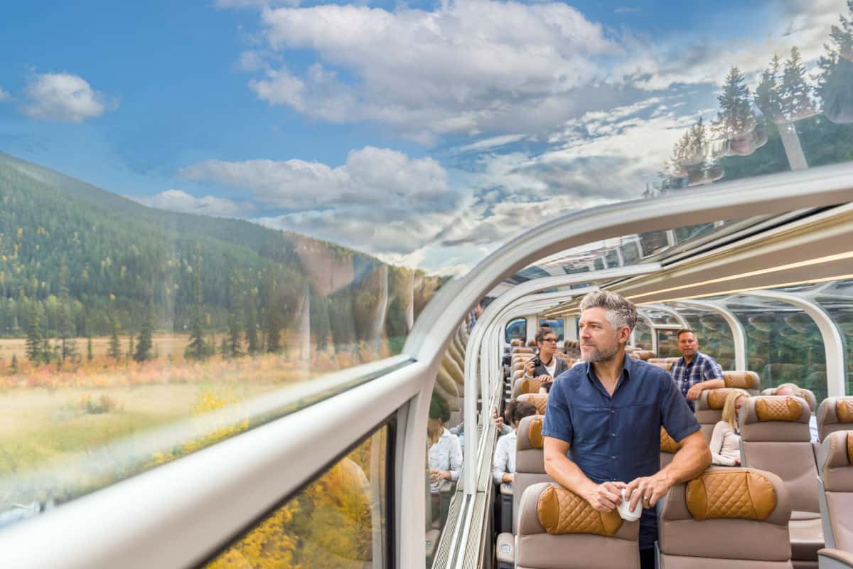 Ein Passagier in einem modernen Zug mit Panoramafenstern, der die Aussicht auf eine grüne Landschaft und den blauen Himmel genießt.