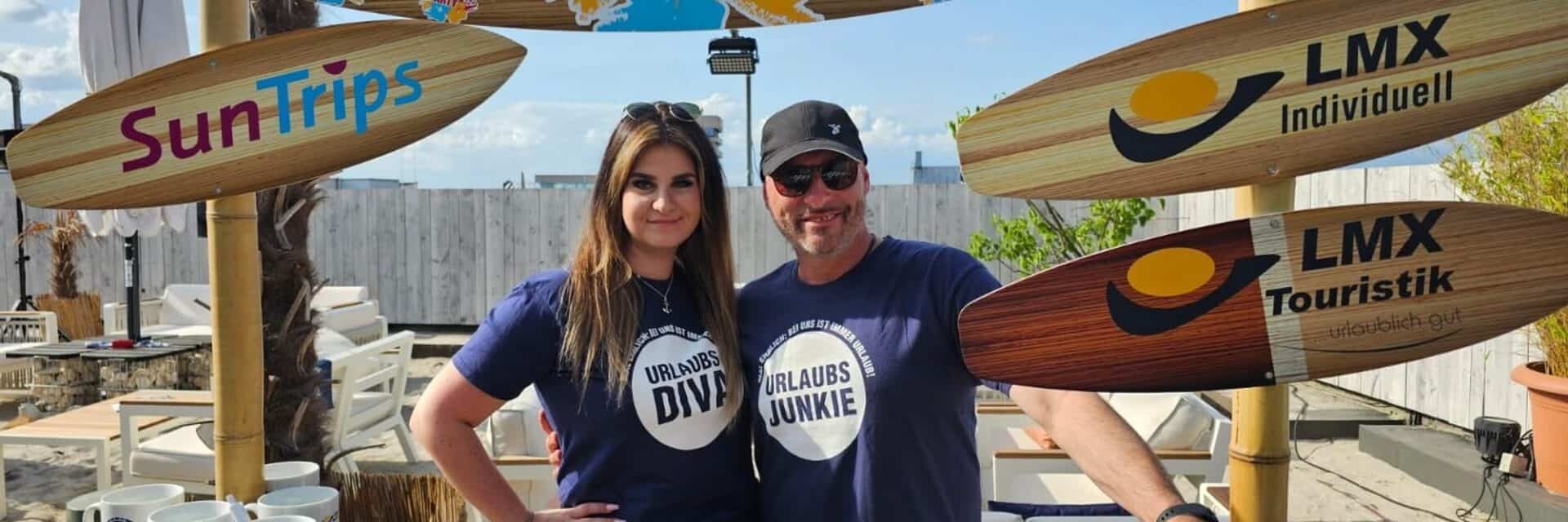 lmx-uebernahme-auctus-krug Zwei Personen stehen vor einem Stand mit Surfbrett-Schilden, die die Marken SunTrips und LMX zeigen. Sie tragen T-Shirts mit Aufdrucken.