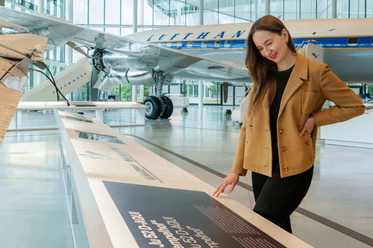 Eine Frau in einem beigen Blazer steht in einem Museum vor einer Informationstafel und betrachtet im Hintergrund ein historisches Lufthansa-Flugzeug.