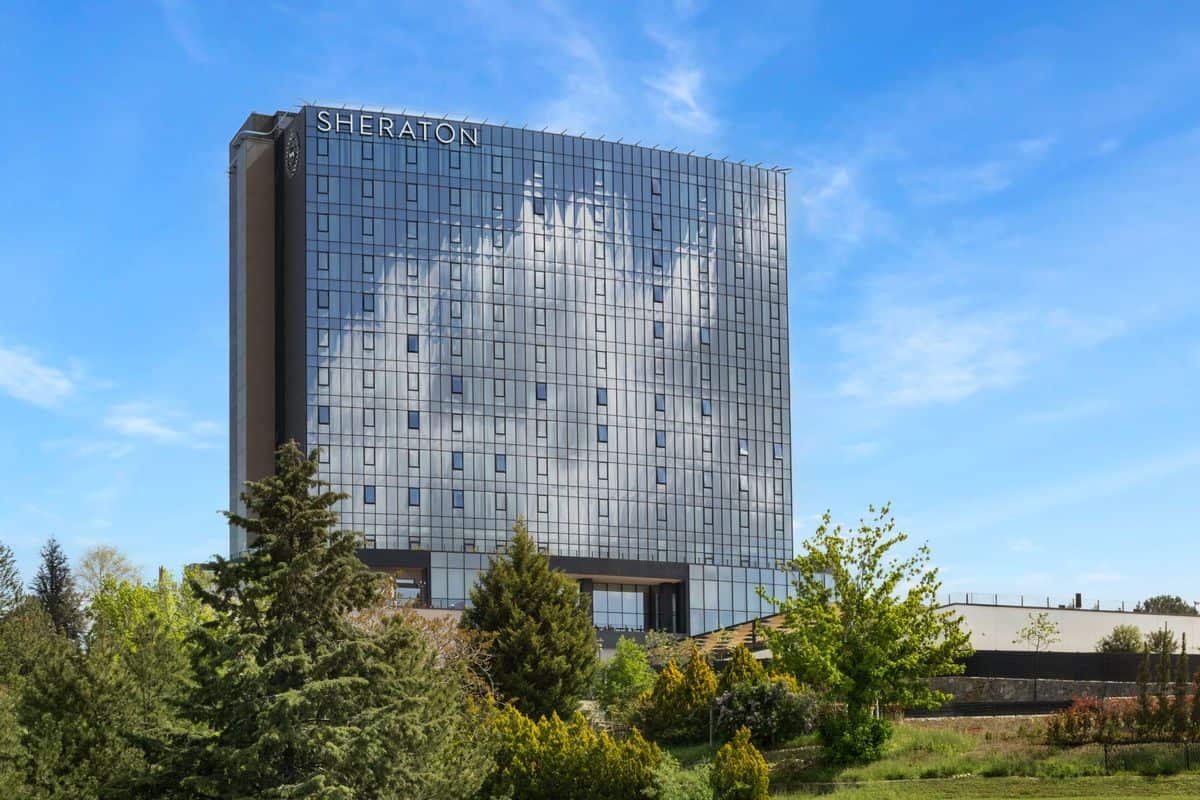 Das moderne Sheraton-Hotel mit reflektierender Glasfassade und umgebenden Bäumen unter blauem Himmel.