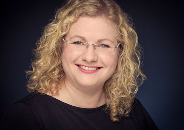 Porträt einer lächelnden Frau mit lockigem, blondem Haar und Brille, vor einem dunklen Hintergrund.