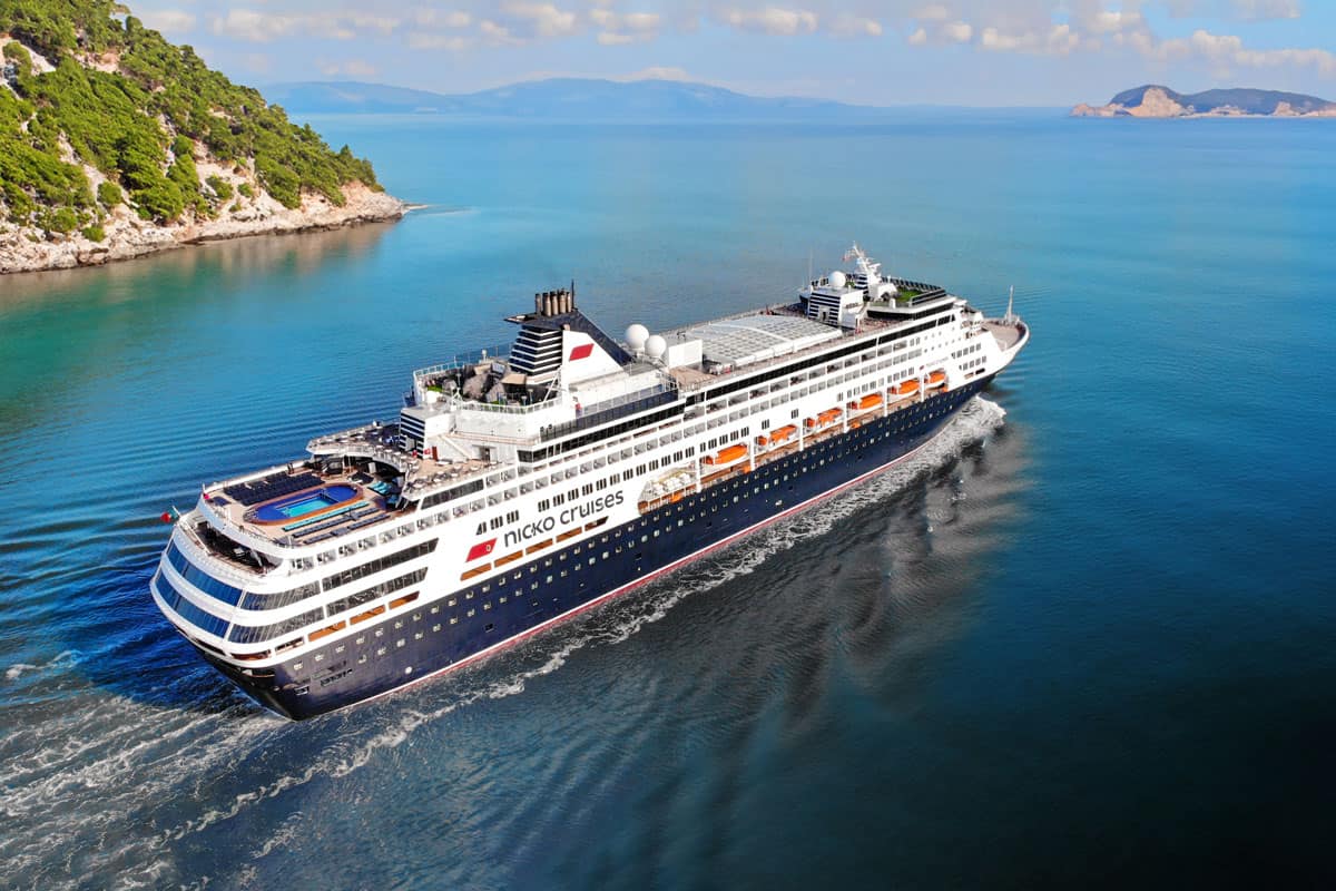 Ein Kreuzfahrtschiff von Nicko Cruises fährt durch ruhiges, blaues Wasser, umgeben von grünen Hügeln und einer klaren, sonnigen Himmel.