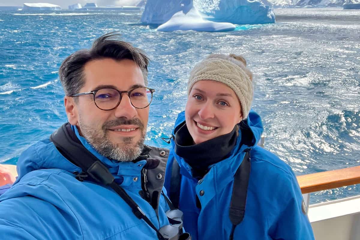 Ein Paar in blauen Jacken posiert auf einem Schiff vor einer beeindruckenden Eisberglandschaft im Ozean.