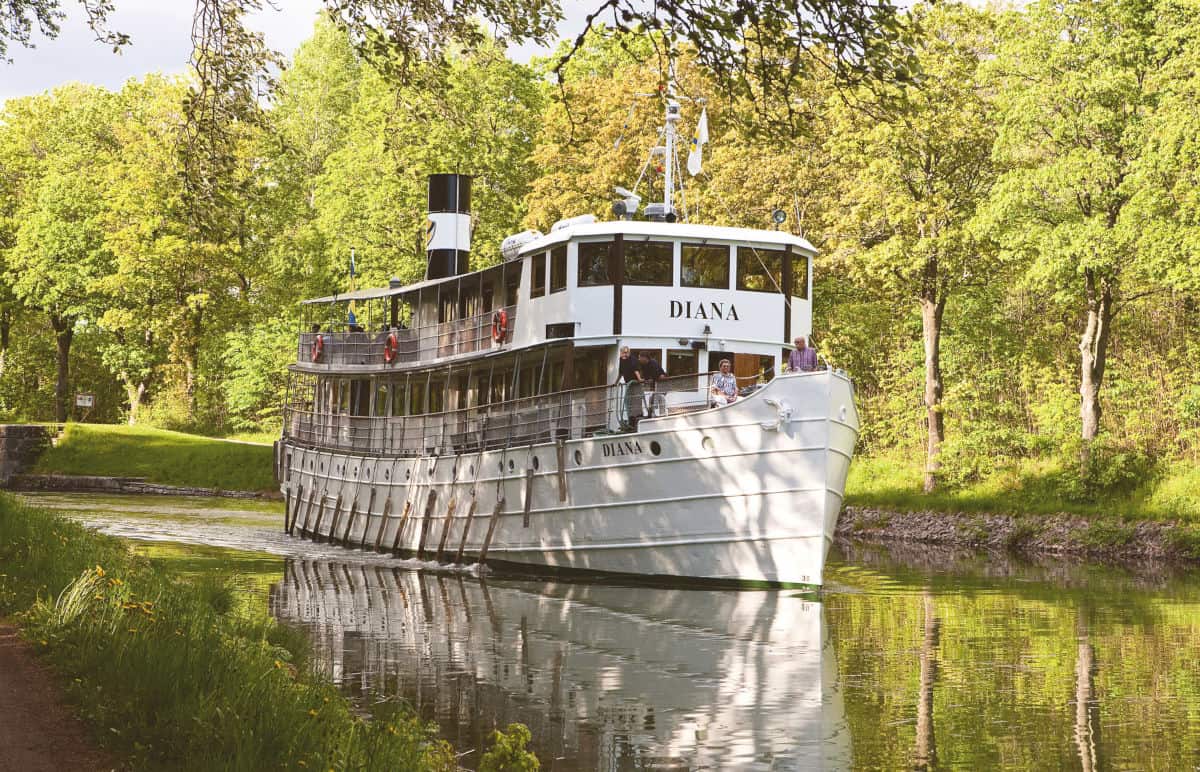 Ein weißes Passagierschiff mit dem Namen Diana fährt auf einem ruhigen Kanal, umgeben von grünen Bäumen und reflektiert im Wasser.