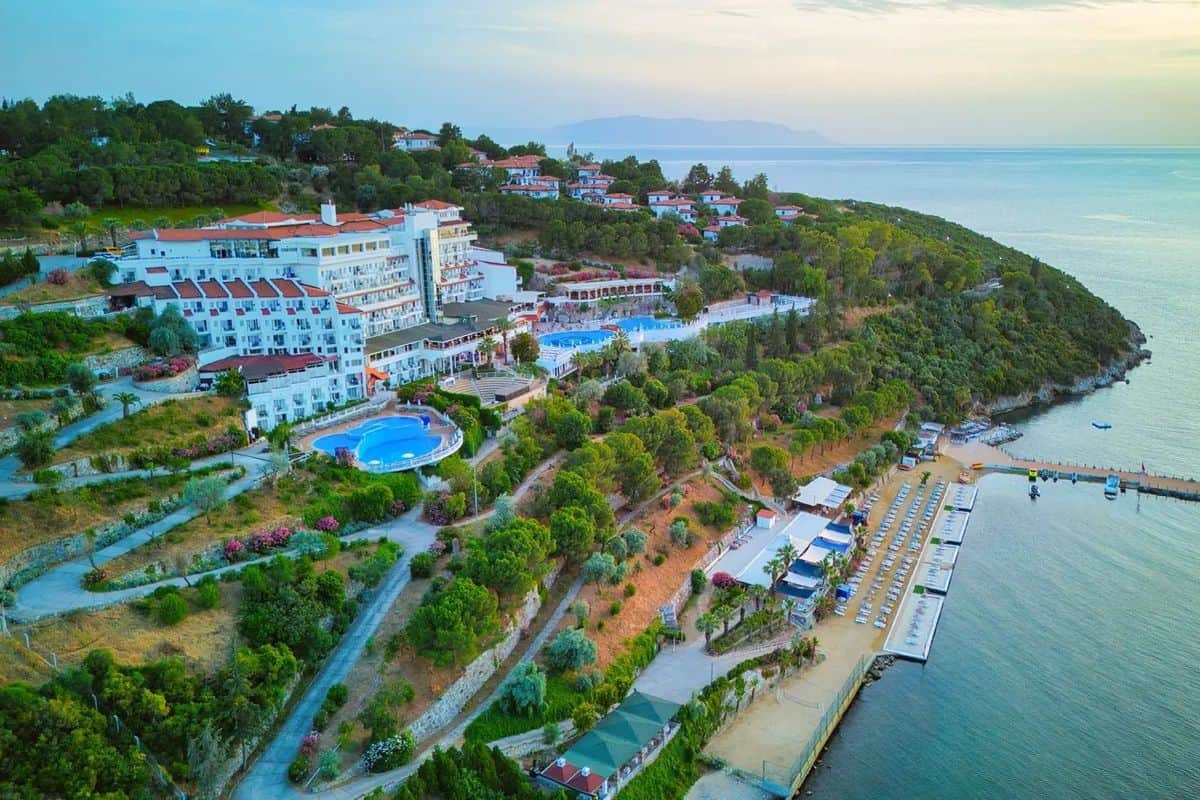 Luftaufnahme eines Resorts an der Küste mit Pools, Gärten und einem Strandbereich, umgeben von Bäumen und dem Meer im Hintergrund.