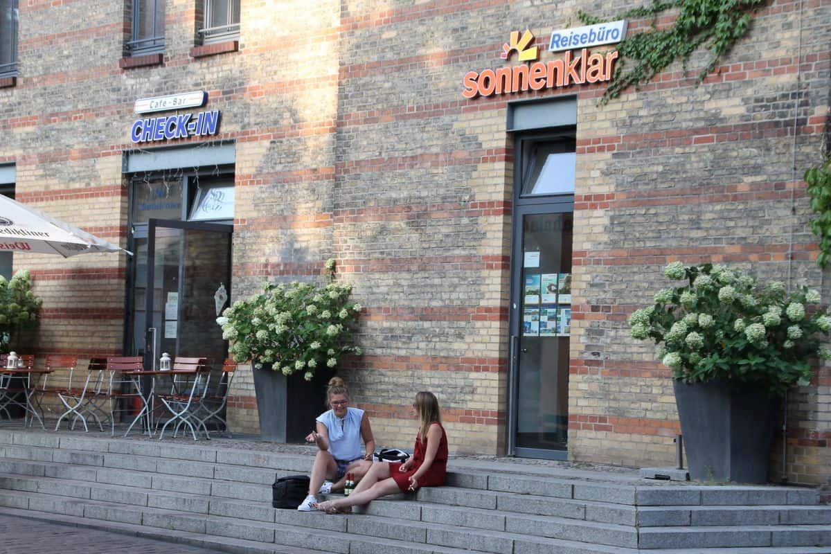 Zwei Frauen sitzen auf einer Treppe vor einem Reisebüro und einem Café, umgeben von großen Pflanzen und einem einladenden Ambiente.
