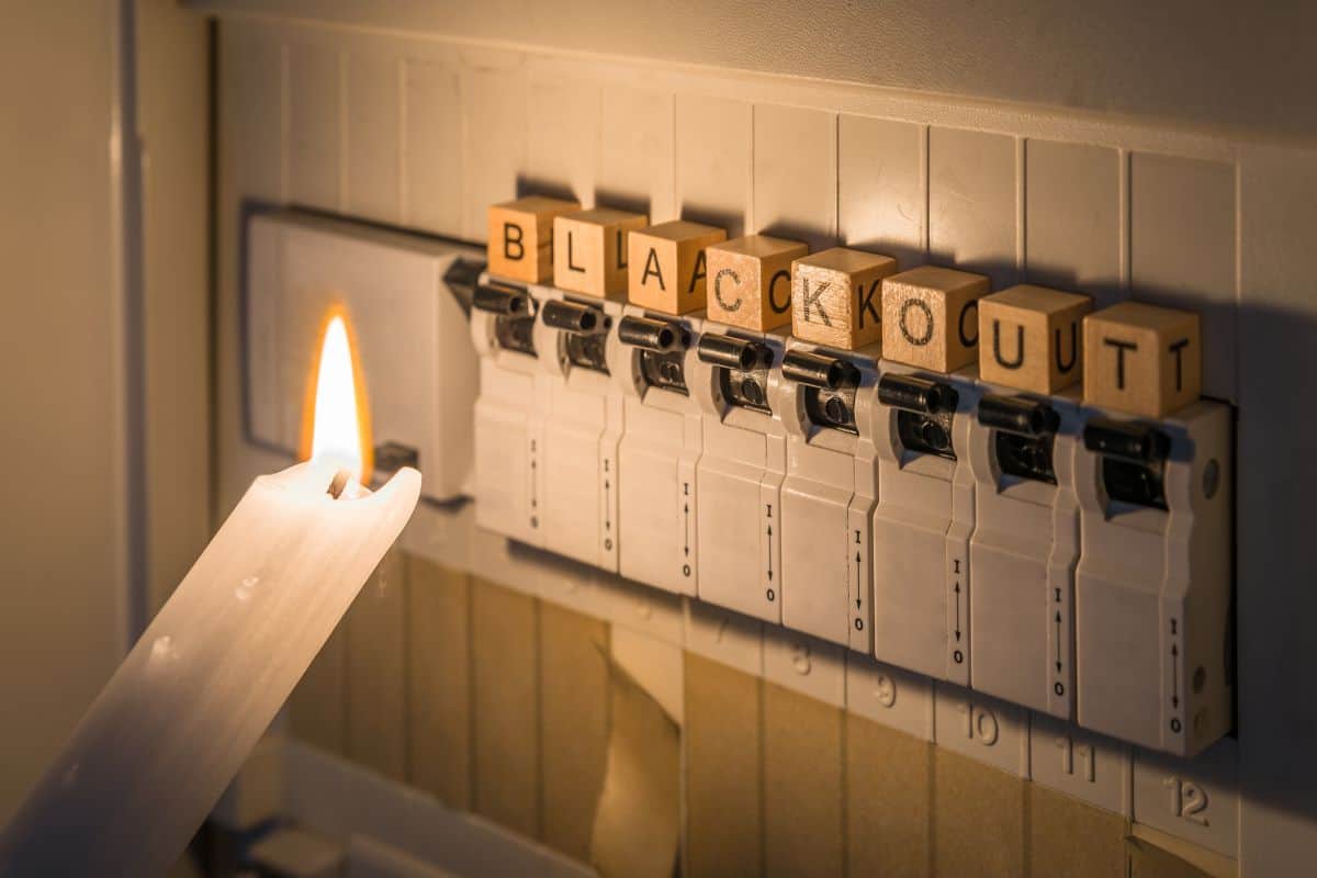Eine brennende Kerze beleuchtet einen Sicherungskasten, auf dessen Schaltern Holzklötze mit den Buchstaben des Wortes BLACKOUT in einer Reihe angeordnet sind.