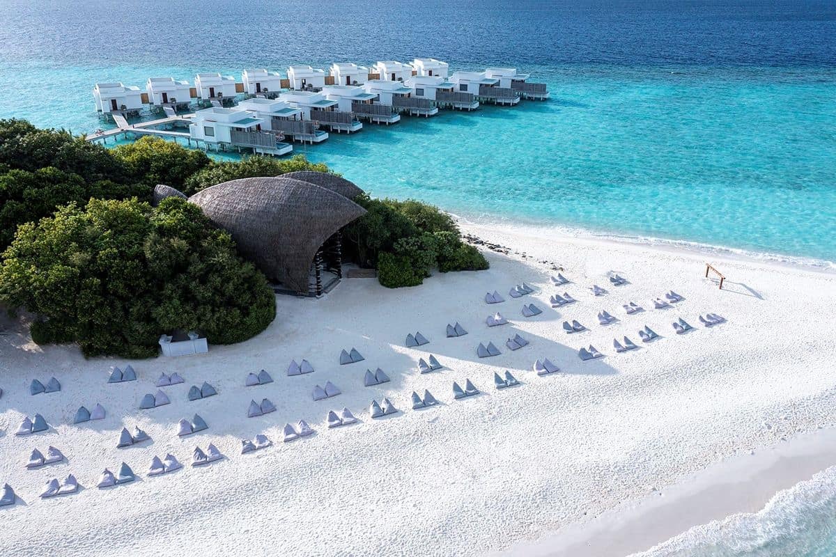 Luftaufnahme eines weißen Sandstrandes mit Liegen und einem modernen Resort im Hintergrund, umgeben von klarem, türkisfarbenem Wasser.