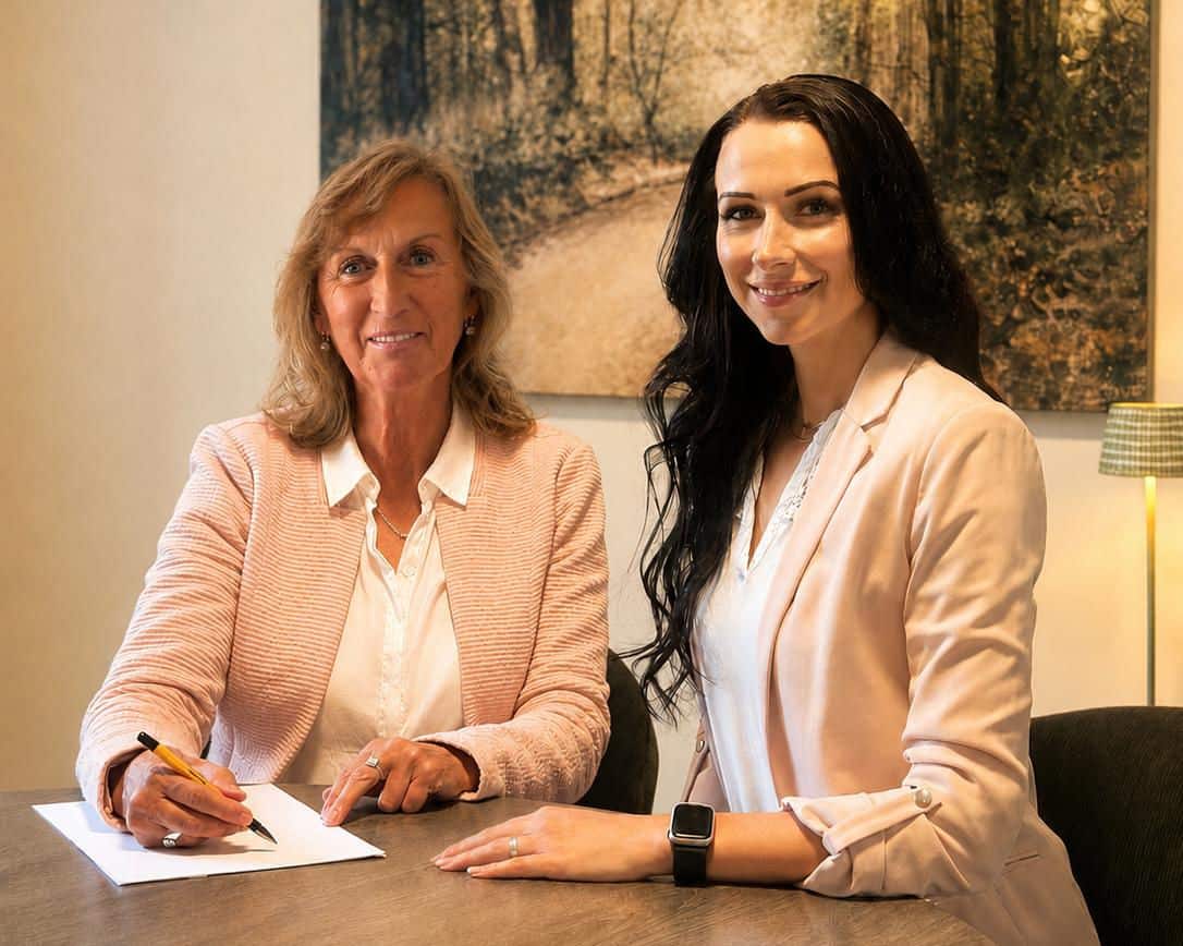 Zwei Frauen in hellen Blazern sitzen an einem Holztisch. Die linke Frau schreibt mit einem Stift auf ein Blatt Papier, während die rechte Frau in die Kamera lächelt.