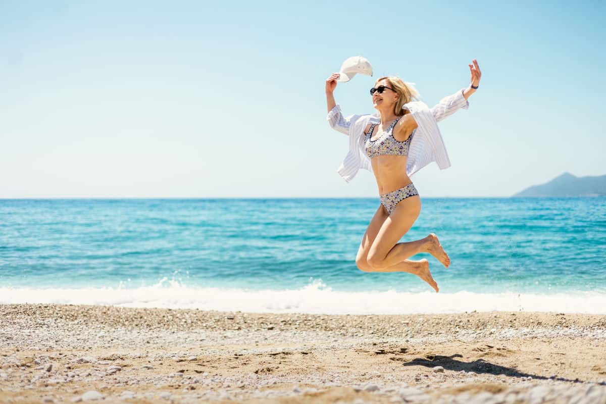 Eine Frau springt fröhlich am Strand, trägt einen Bikini und ein offenes Hemd, mit dem Meer im Hintergrund und klarem Himmel.