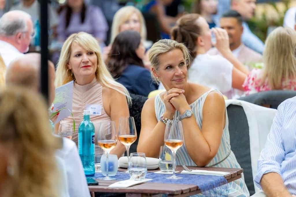 Zwei blonde Frauen sitzen an einem gedeckten Tisch im Freien, vor ihnen stehen Weingläser und eine Wasserflasche, im Hintergrund weitere Gäste bei Tageslicht.
