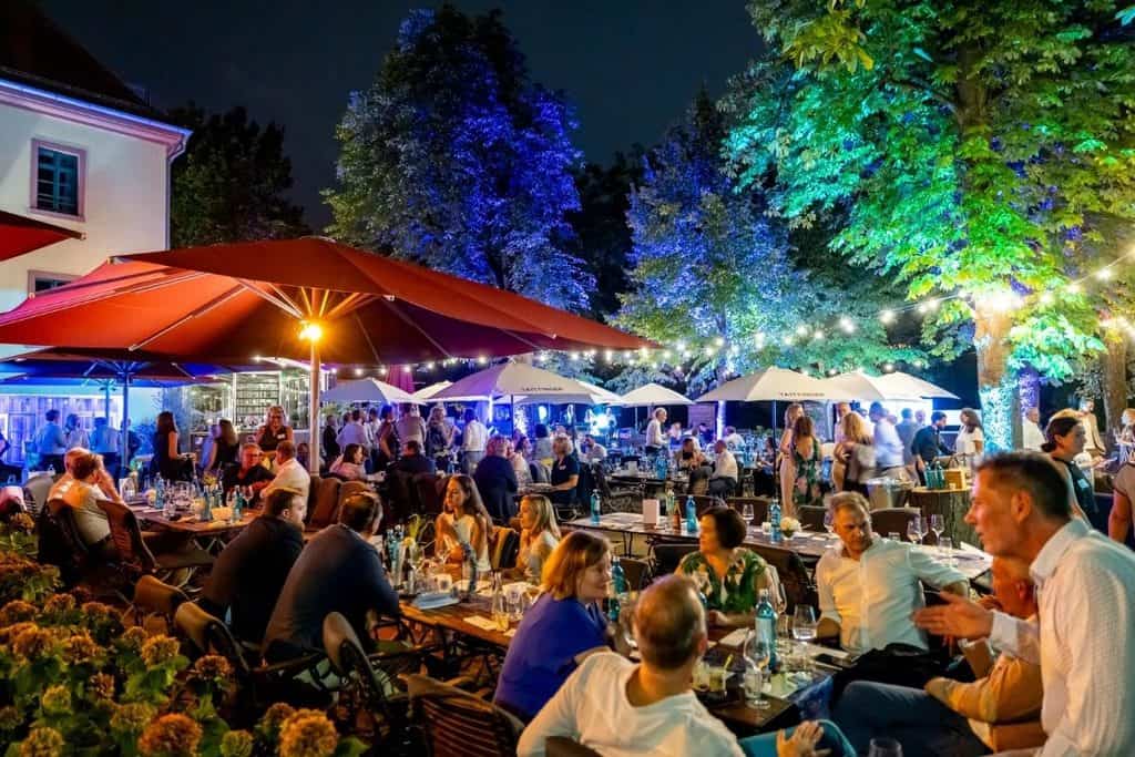 Eine abendliche Außenveranstaltung mit vielen Gästen an langen Tischen unter großen Sonnenschirmen, beleuchtet durch farbige Strahler und Lichterketten.