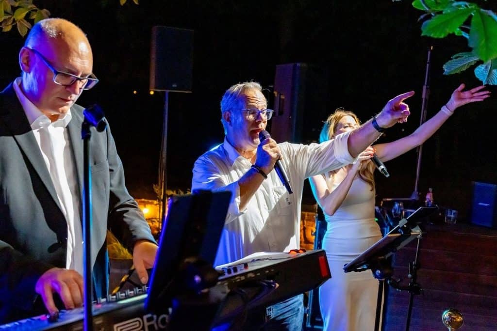 Drei Personen auf einer Bühne bei Nacht: Ein Mann am Keyboard, ein singender Mann mit Mikrofon und eine Frau, die mit ausgestrecktem Arm singt.