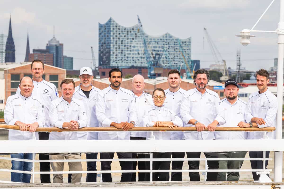 Eine Gruppe von zehn Personen in weißen Kochjacken steht auf einem Schiffsdeck vor der Kulisse der Hamburger Elbphilharmonie bei hellem Tageslicht.