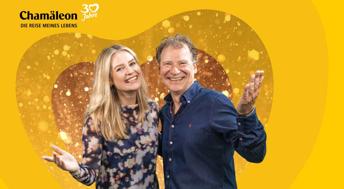 Eine Frau mit blondem Haar und ein Mann mit kurzem Haar lächeln vor einem herzförmigen, gold glitzernden Hintergrund mit dem Chamäleon-Logo in der Ecke.