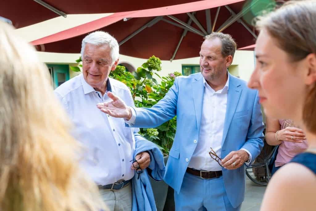 Zwei Männer im Gespräch unter einem roten Sonnenschirm im Freien, einer trägt ein hellblaues Sakko und gestikuliert, der andere ein weißes Hemd mit Brille.