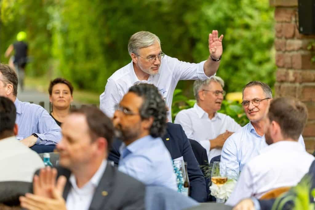 Ein Mann mit Brille und weißem Hemd steht in einer Gruppe von Menschen im Freien auf und hebt eine Hand, während andere Personen an Tischen sitzen.